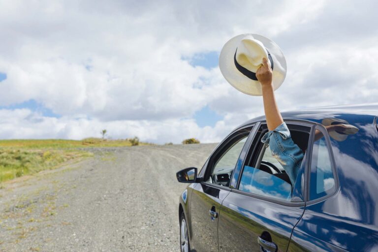 Nell'immagine si vede un' auto blu su una strada sterrata. dall'auto esce il braccio di una ragazza con in mano un cappello.