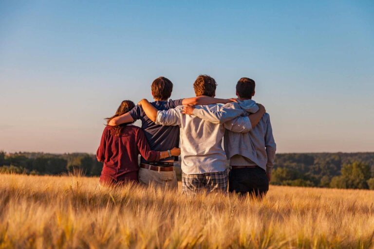 Immagine di quattro giovani ragazzi abbracciati in mezzo ad un campo. I ragazzi sono uno affianco all'altro che fissano l'orizzonte.